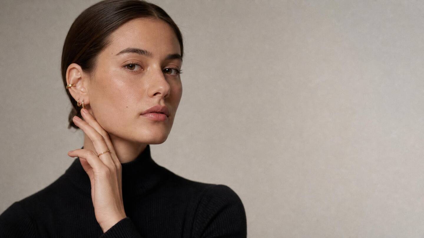 A woman in a black roll-neck, hand raised to her jaw, wearing a fine gold ear cuff, huggie hoops and a slim chain ring